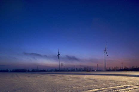 Zwei Windkraftanlagen heben sich bei Sonnenuntergang gegen den klaren Himmel ab. Der Boden ist mit Schnee bedeckt.