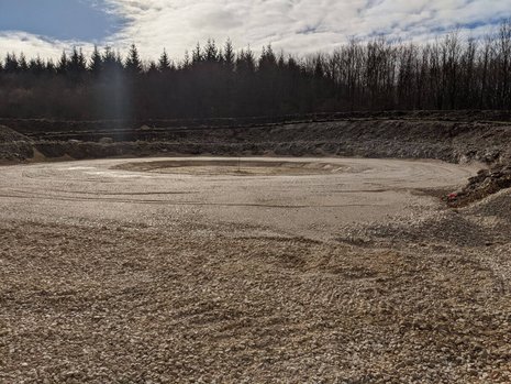 Eine große Vertiefung in der Erde. Die Erde wurde kreisförmig an den Rand geschoben. In der Mitte ist ein kreisförmiges Fundament entstanden.