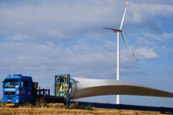 Ein Rotorblatt einer Windanlage liegt auf einem LKW. Im Hintergrund steht eine bereits fertige Windanlage.