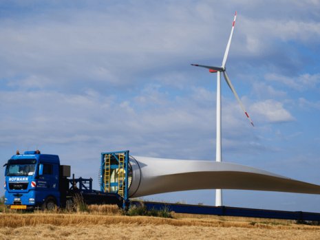 Ein Rotorblatt einer Windanlage liegt auf einem LKW. Im Hintergrund steht eine bereits fertige Windanlage.