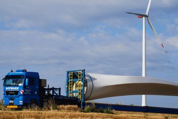 Ein Rotorblatt einer Windanlage liegt auf einem LKW. Im Hintergrund steht eine bereits fertige Windanlage.