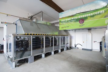 Links ein Windgas-Elektrolyseur, rechts ein Banner mit der Aufschrift "Aus Wind wird Wasserstoff!".