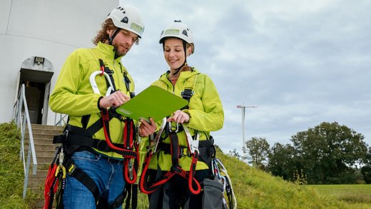 Zwei Personen mit Arbeitsgurten und Helmen schauen auf ein Klemmbrett. Hinter ihnen zwei Windräder.