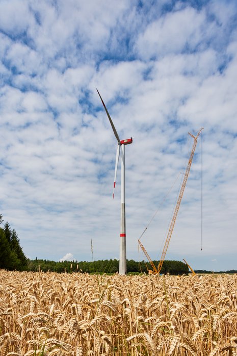 Windkraftanlage in der Mitte mit einem gelben Kran auf der rechten Seite und einem teilweise bewölkten Himmel im Hintergrund.