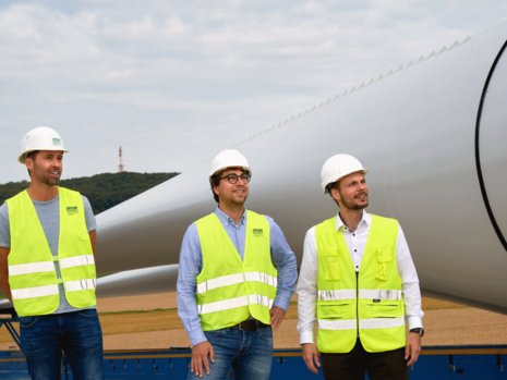 Drei Arbeiter mit Bauhelmen und Warnwesten schauen hoch zu einer Windanlage. Im Hintergrund liegt ein Rotorblatt einer Windanlage.