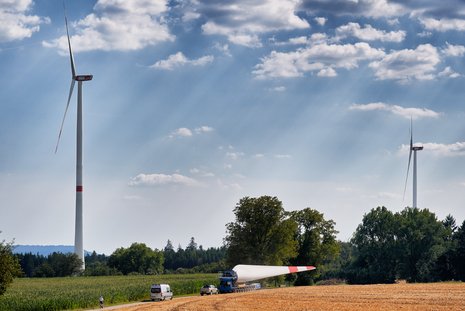 Ein Fahrzeug transportiert ein Windradflügel und im Hintergrund tauchen zwei Windräder auf.