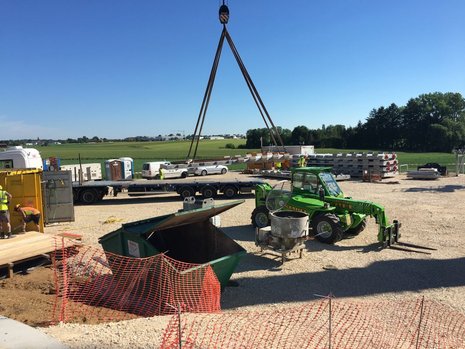 Baustelle mit einem gelben Container auf der linken Seite, einer grünen Tonne in der Mitte, einem grünen Bagger auf der rechten Seite und einem Baufahrzeug im Hintergrund.