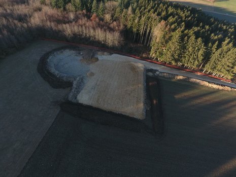 Eine kreisförmige Vertiefung in der Erde aus der Vogelperspektive. Die Erde wurde an den Rand geschoben. An dieses grenzt ein umgegrabenes quadratisches Feld.