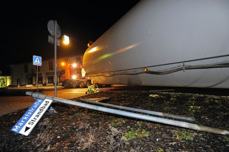 ein Windradflügel, der nachts durch eine Dorfstraße transportiert wird. Zwei Straßenschilder sind entwurzelt und liegen im Vordergrund.