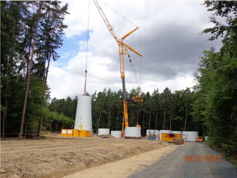 Kran hebt einen Teil des Mastes der Windkraftanlage an. Im Hintergrund ein Wald.