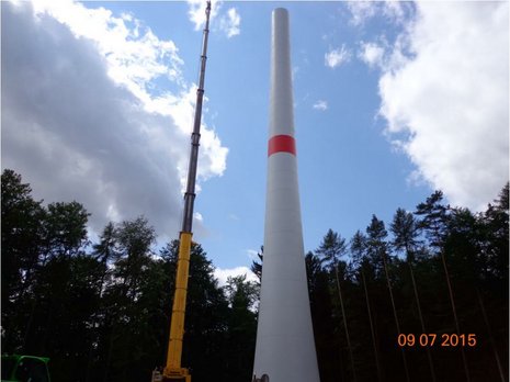 Blick von unten auf einen Kran, der einen Windkraftanlagenmast montiert. Im Hintergrund ein Wald.