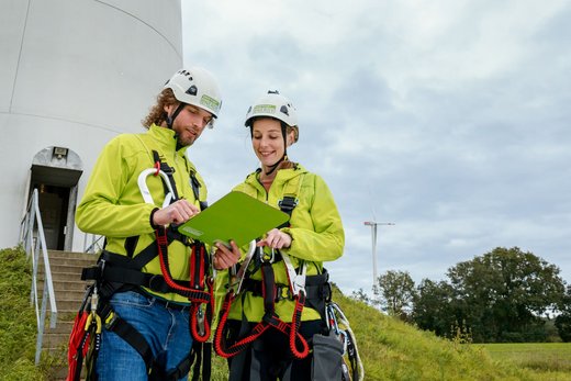 Zwei Personen mit Arbeitsgurten und Helmen schauen auf ein Klemmbrett. Hinter ihnen zwei Windräder.