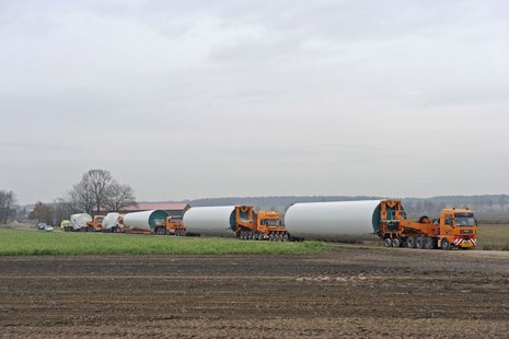 4 Lastwagen transportieren 3 Teile des Mastes der Windkraftanlage durch ein Feld.