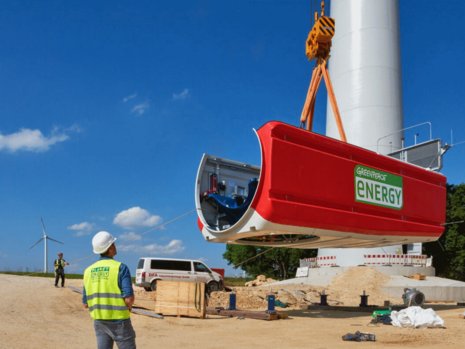 Ein rotes Windturbinenmodul wird von einem Kran angehoben. Personen in Sicherheitswesten beobachten den Vorgang auf einer Baustelle mit Windrädern und Fahrzeugen. Text auf Modul: "GREENPEACE ENERGY".