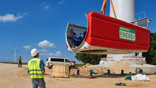 Ein rotes Windturbinenmodul wird von einem Kran angehoben. Personen in Sicherheitswesten beobachten den Vorgang auf einer Baustelle mit Windrädern und Fahrzeugen. Text auf Modul: "GREENPEACE ENERGY".