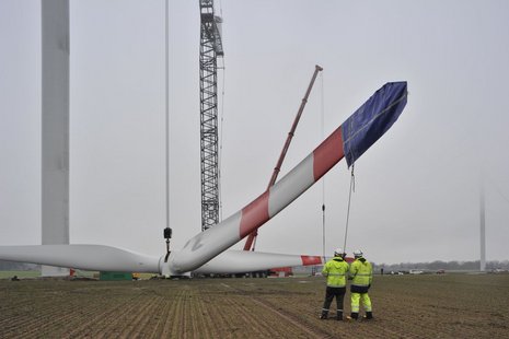 Die 3 Flügel der Windkraftanlage werden von einem Kran angehoben, während 2 Arbeiter den Vorgang überwachen.