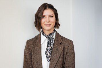 Eine junge Frau mit braunen kinnlangen Haaren blickt freundlich in die Kamera. Sie trägt ein gebundenes Halstuch und einen braunen Stoff-Blazer.