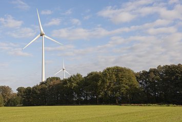Zwei Windkraftanlagen stehen am Rand einer grünen Wiese vor einem Baumsaum unter leicht bewölktem Himmel.
