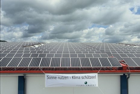 Banner mit Aufschrift "Sonne nutzen - Klima schützen!" an Gebäude mit zahlreichen Solarmodulen auf dem Dach vor bewölktem Himmel