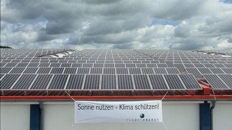 Banner mit Aufschrift "Sonne nutzen - Klima schützen!" an Gebäude mit zahlreichen Solarmodulen auf dem Dach vor bewölktem Himmel