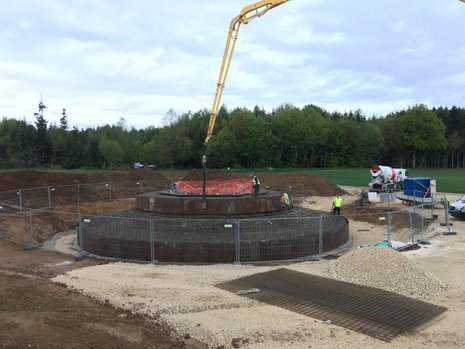 Rundes Erdloch mit einem umgebenden Zaun und 3 Arbeitern und einem Baufahrzeug.