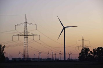Windkraftanlage und Strommasten in einer Landschaft bei Sonnenuntergang, mit mehreren Windrädern im Hintergrund und sichtbaren Hochspannungsleitungen.